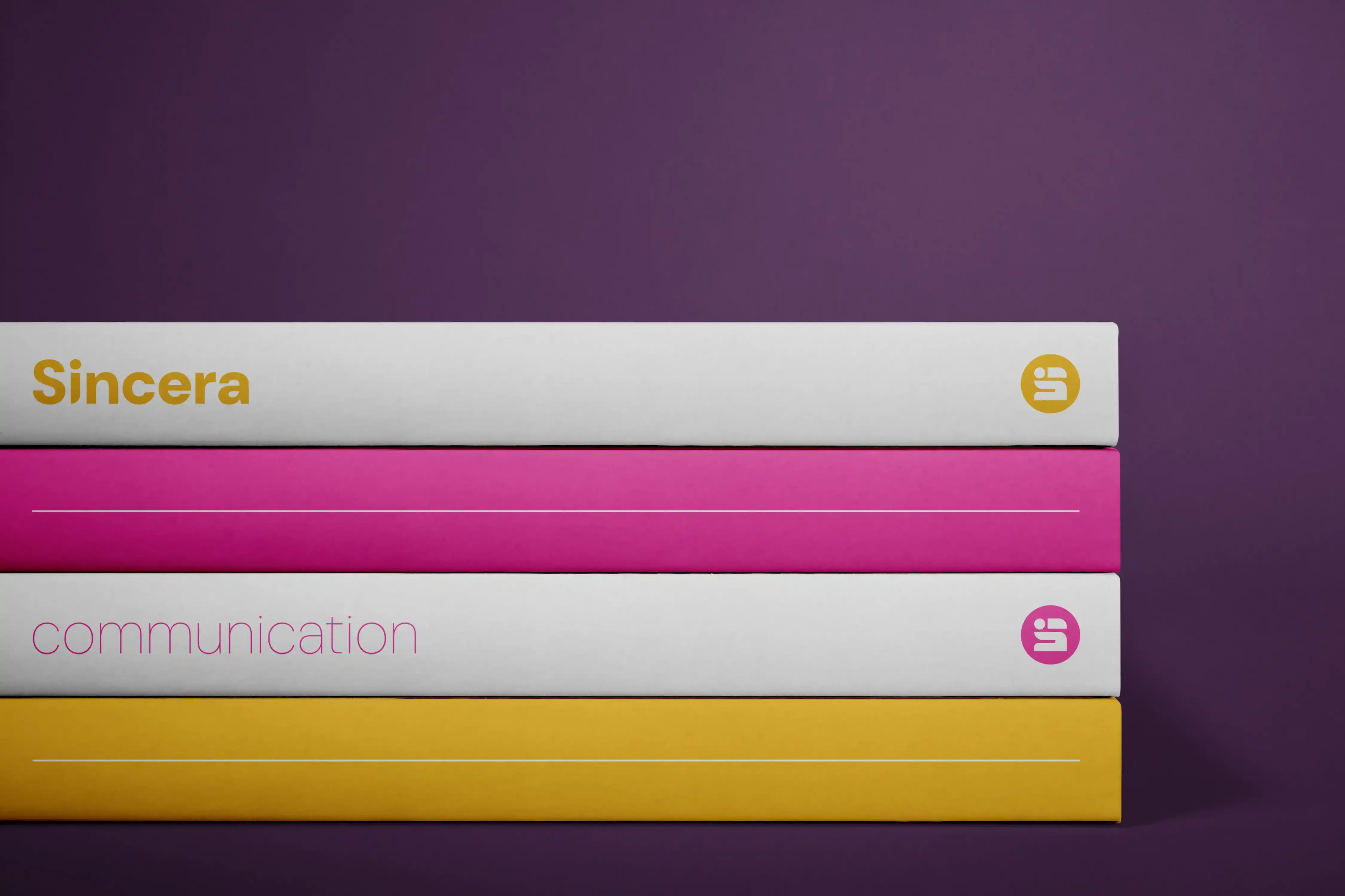 Photo de quatre livre empilés. Le premier est blanc et à comme titre : 'Sincera' écrit en jaune avec l'icône de la marque en blanc dans un cercle jaune placée à droite de la tranche. Le deuxième livre est rose. Le troisième est blanc avec comme titre : 'communication' écrit en rose avec l'icône de la marque en blanc dans un cercle rose placée à droite de la tranche. Le quatrième livre est jaune.